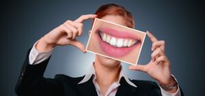 ALT TEXT: Woman holding picture of a smile. IMAGE URL: https://pixabay.com/photos/woman-smile-teeth-health-tooth-3498849/ 